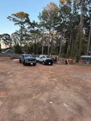 Two pickup trucks parked on a cleared red-clay lot ringed by tall pines