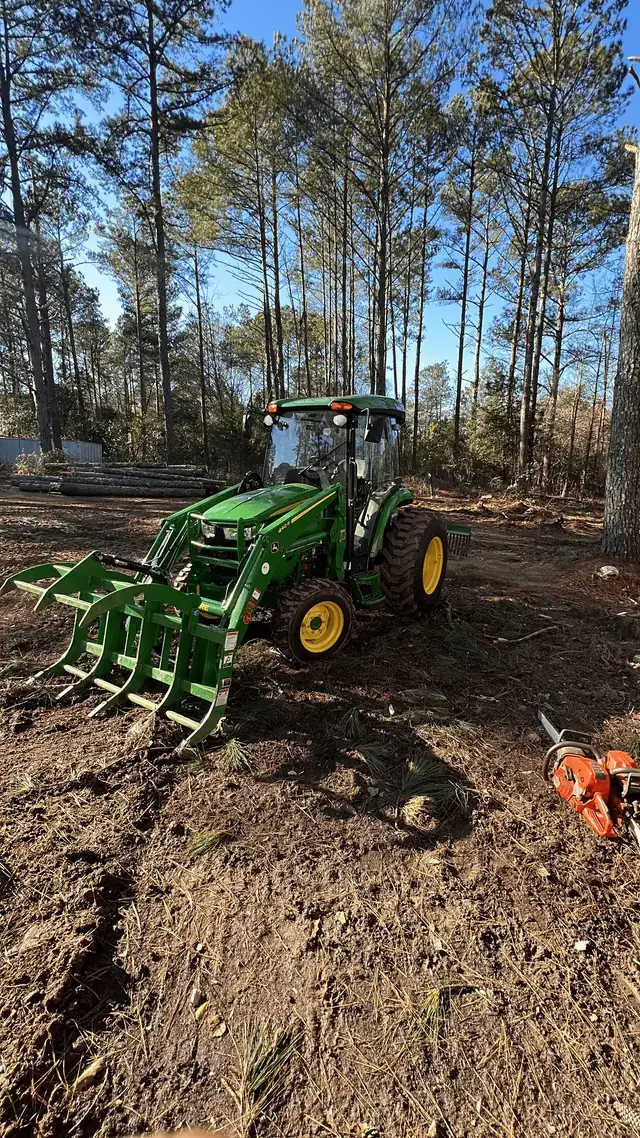Green tractor with a grapple attachment on a freshly cleared lot among pines