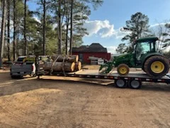 Green tractor on a flatbed trailer beside a load of cut log sections